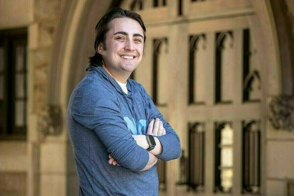 Smiling student wearing a blue sweatshirt stands with arms crossed in front of an arched doorway on campus.